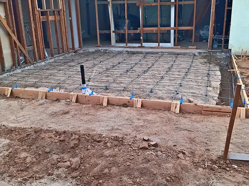 Formed slab with rebar grid and plumbing stub-up in front of a stud-framed home addition, ready for concrete pour