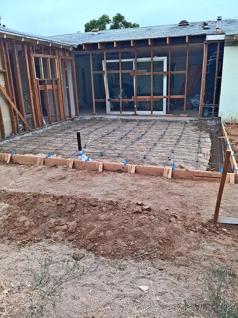Formed slab with rebar grid and plumbing stub-up in front of a stud-framed home addition, ready for concrete pour