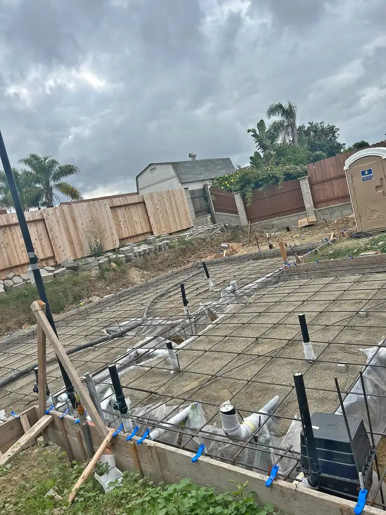 Rebar grid and plumbing stubs set over gravel inside wood forms for a backyard concrete slab before pouring