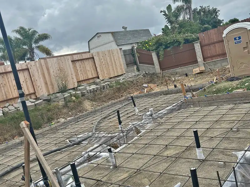 Rebar grid and plumbing stubs set over gravel inside wood forms for a backyard concrete slab before pouring