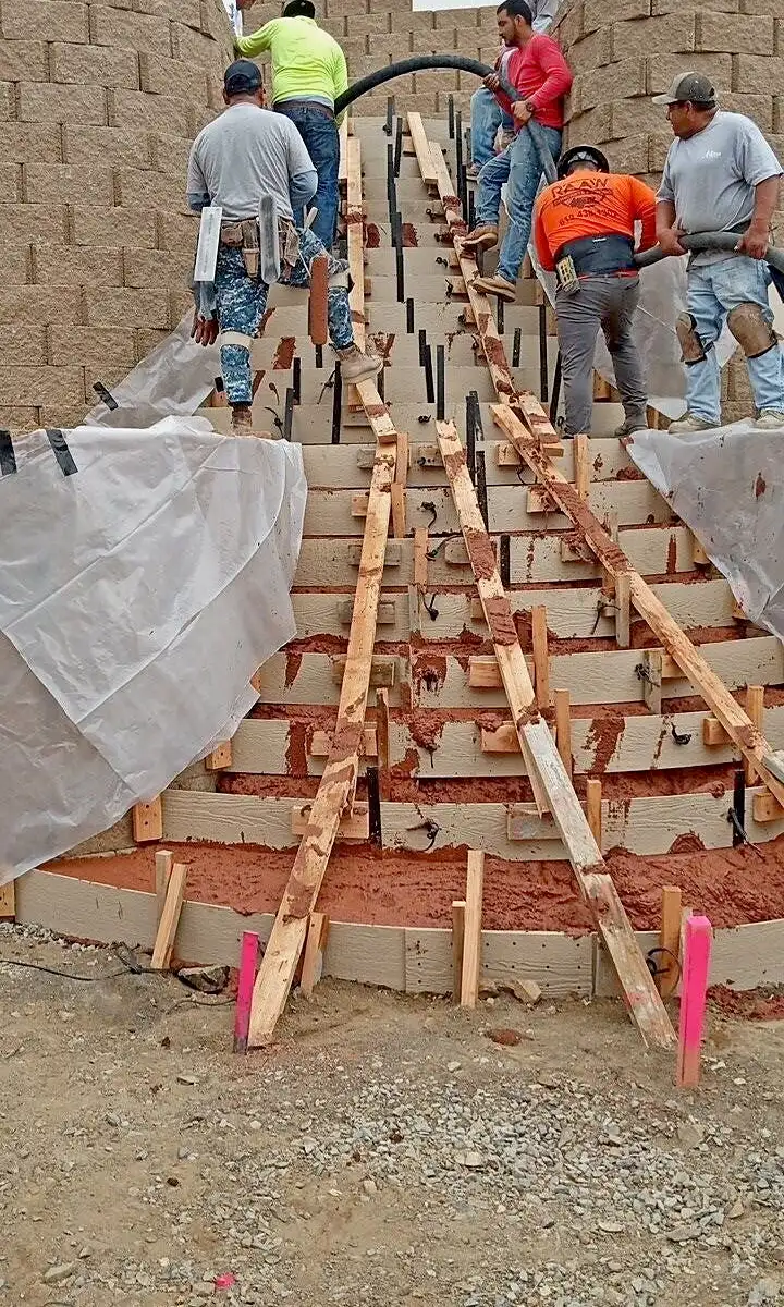 Crew pouring a curved concrete staircase through step forms between two block retaining walls