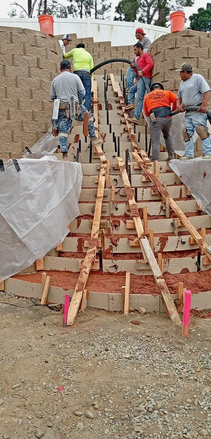Crew pouring a curved concrete staircase through step forms between two block retaining walls