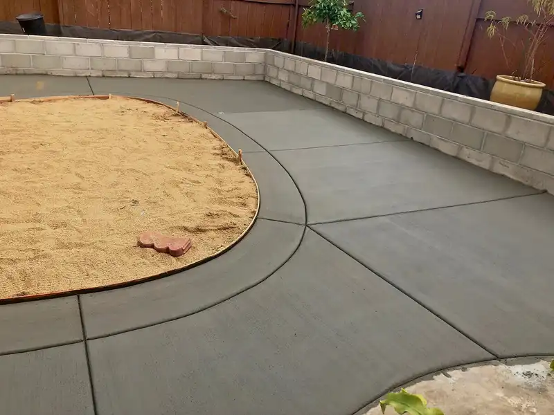 Freshly poured backyard concrete patio with curved border around circular sand area beside a block wall