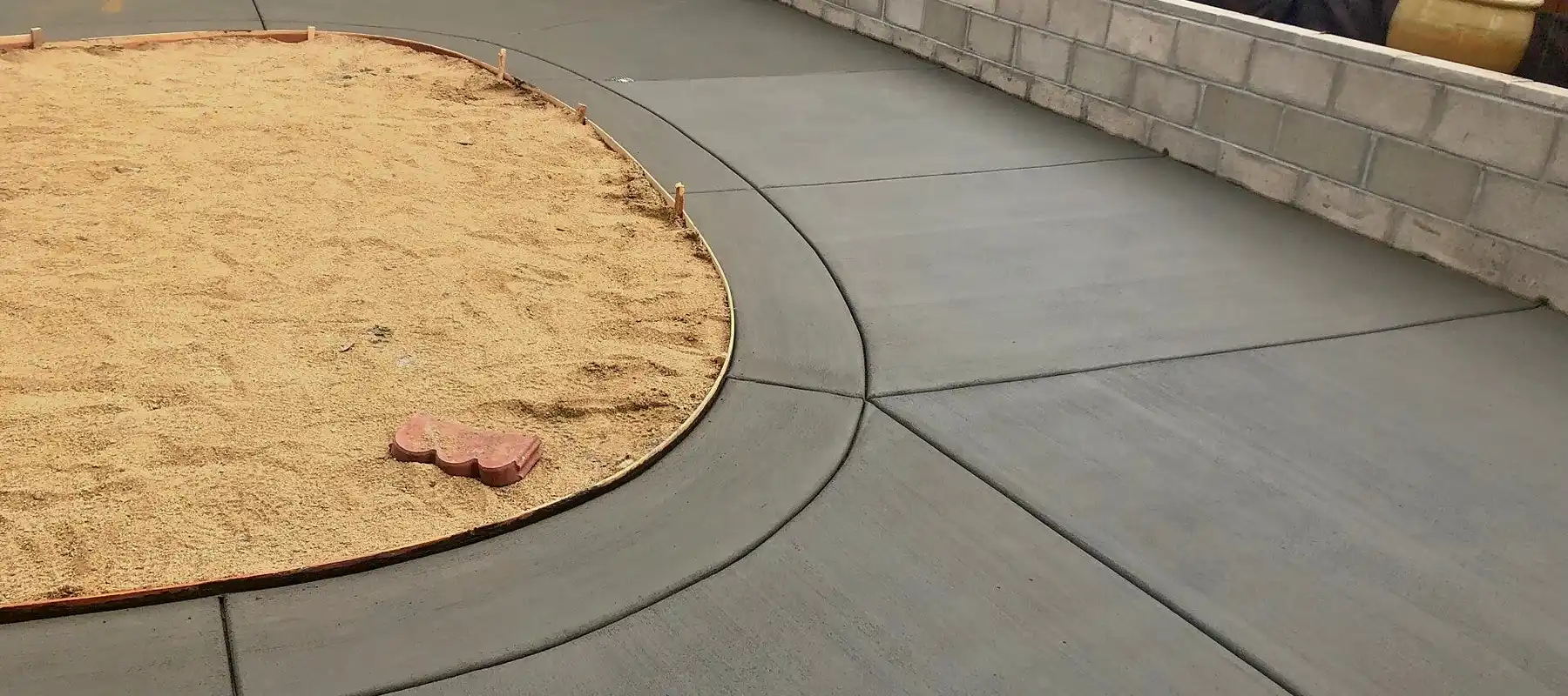Freshly poured backyard concrete patio with curved border around circular sand area beside a block wall