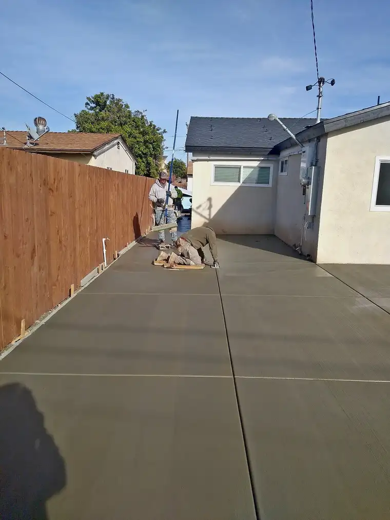 Crew finishing a freshly poured concrete driveway beside a wood fence and stucco home