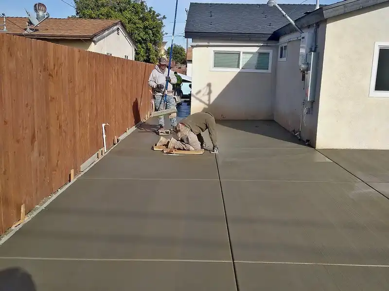 Crew finishing a freshly poured concrete driveway beside a wood fence and stucco home
