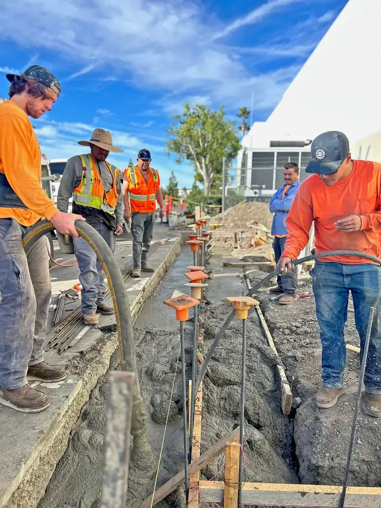 Concrete crew pumping concrete into a rebar-lined footing trench beside a commercial building