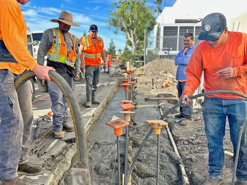 Concrete crew pumping concrete into a rebar-lined footing trench beside a commercial building