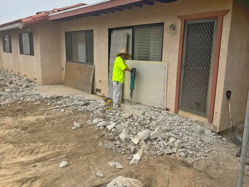 Worker with demolition hammer breaking up old concrete patio behind a stucco home, rubble piled along the wall
