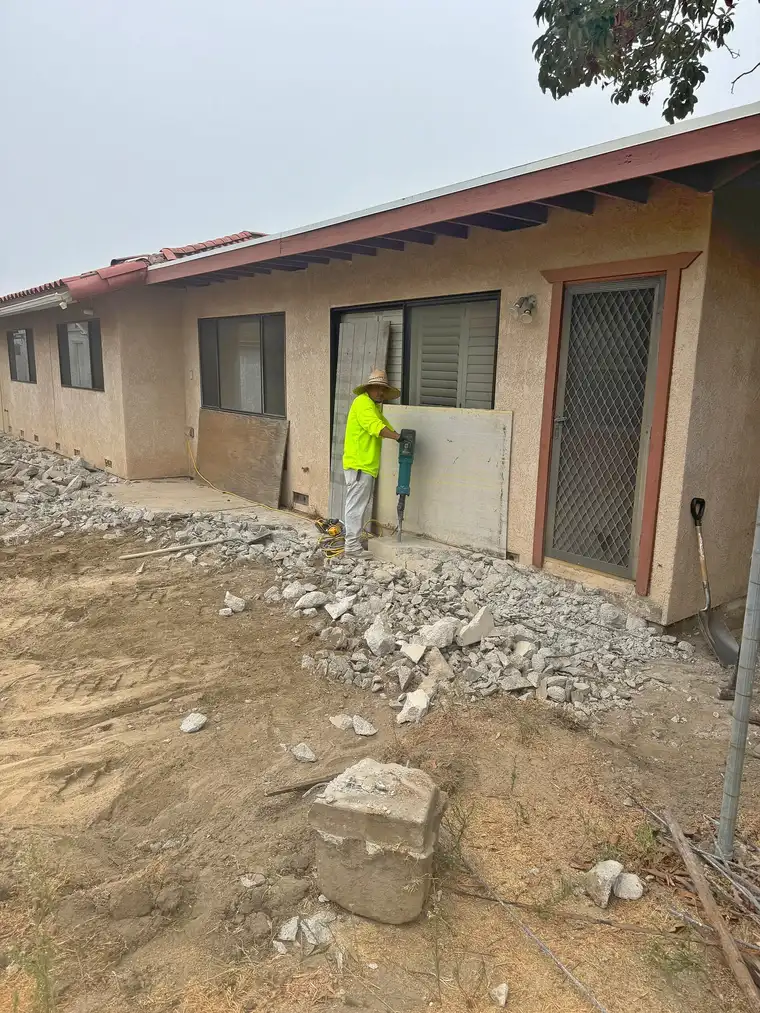 Worker with demolition hammer breaking up old concrete patio behind a stucco home, rubble piled along the wall