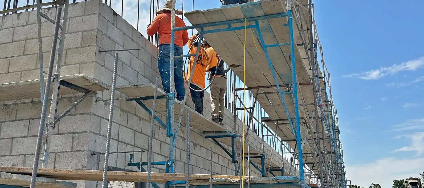 Crew on scaffolding building a tall concrete block wall with rebar projecting from the top