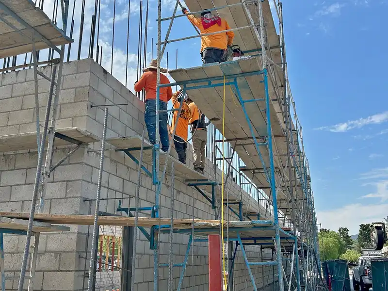 Crew on scaffolding building a tall concrete block wall with rebar projecting from the top