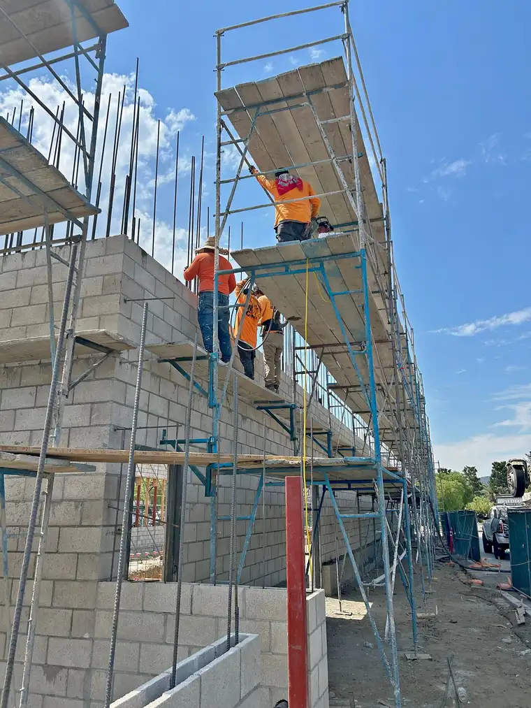 Crew on scaffolding building a tall concrete block wall with rebar projecting from the top